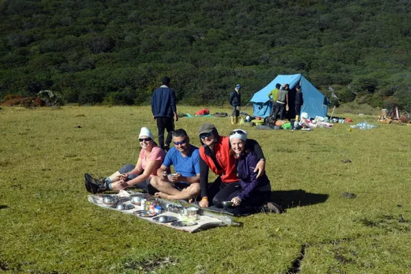 Group of trekkers relaxing and enjoying a camp meal on a grassy mountain meadow near a blue tent.