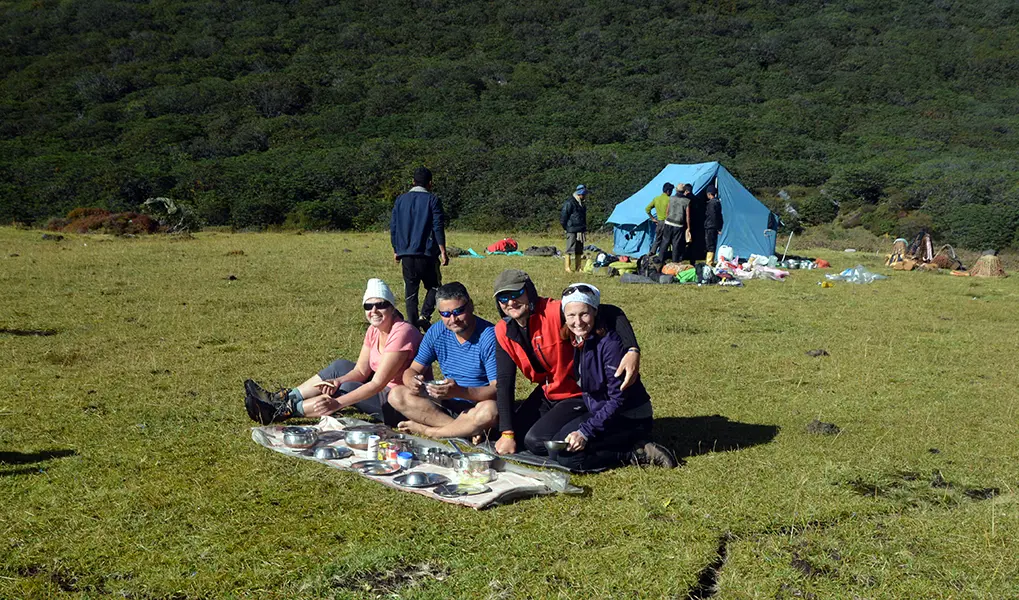 Group of trekkers relaxing and enjoying a camp meal on a grassy mountain meadow near a blue tent.