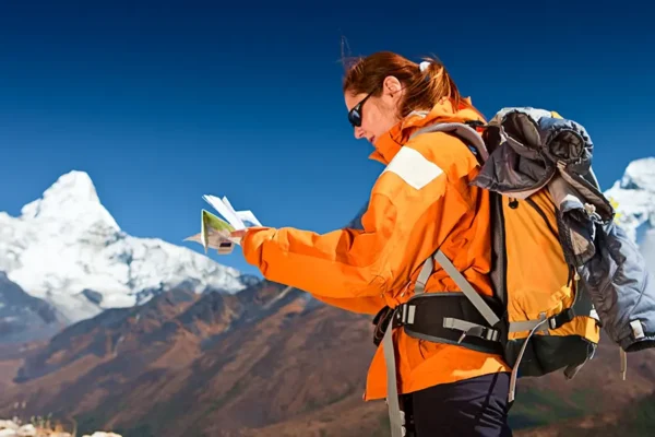 Female trekker with a backpack reading a trail map against a backdrop of snow-capped mountain peaks.
