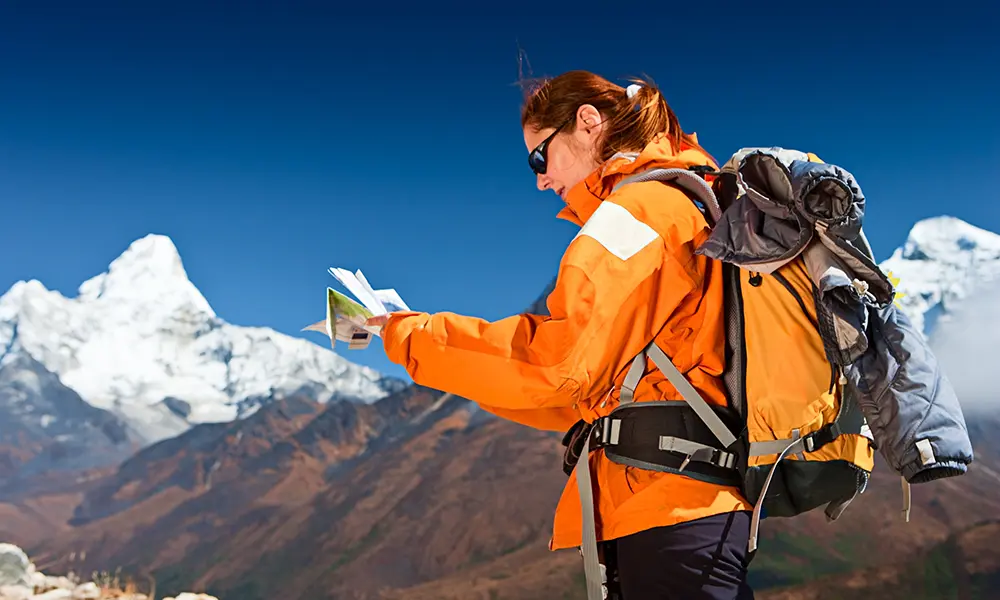 Female trekker with a backpack reading a trail map against a backdrop of snow-capped mountain peaks.