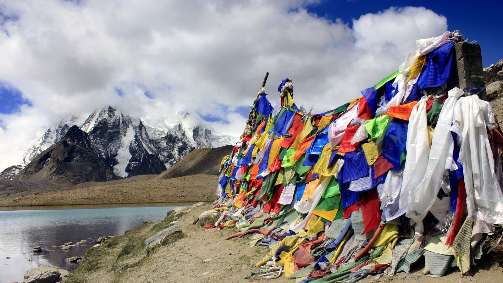 Colorful Buddhist prayer flags beside the turquoise waters of Gurudongmar Lake with snow-capped mountains in North Sikkim, India"