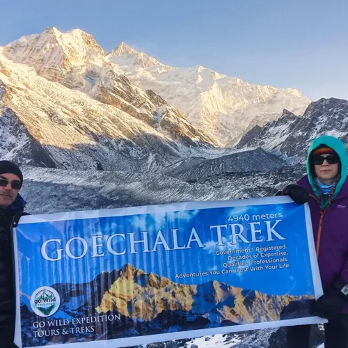 Two trekkers holding a banner at Goechala Trek Viewpoint 1 against the snow-capped Kanchenjunga mountains.