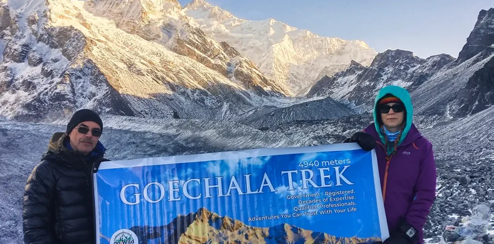 Two trekkers holding a banner at Goechala Trek Viewpoint 1 against the snow-capped Kanchenjunga mountains.