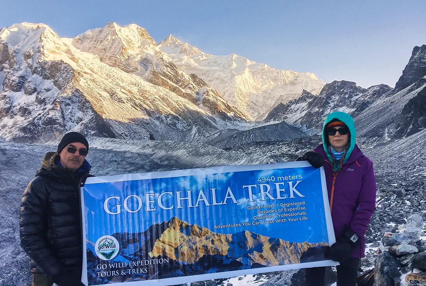 Two trekkers holding a banner at Goechala Trek Viewpoint 1 against the snow-capped Kanchenjunga mountains.