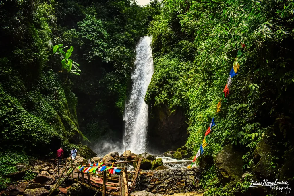 Cascading Kanchenjunga Waterfalls in West Sikkim, featuring a wooden bridge and colorful prayer flags.