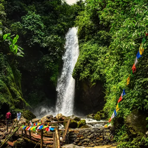 Cascading Kanchenjunga Waterfalls in West Sikkim, featuring a wooden bridge and colorful prayer flags.