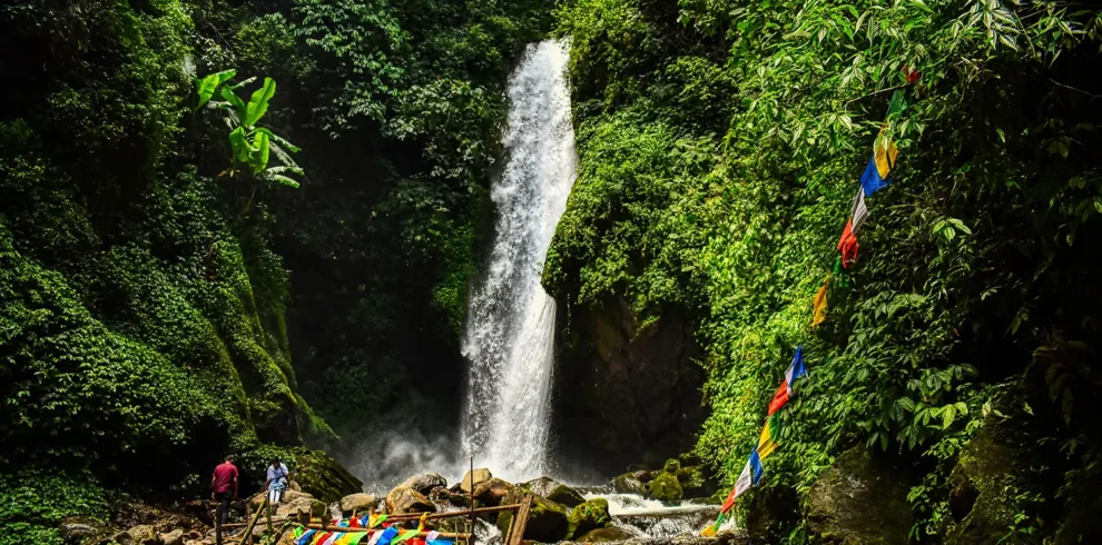Cascading Kanchenjunga Waterfalls in West Sikkim, featuring a wooden bridge and colorful prayer flags.