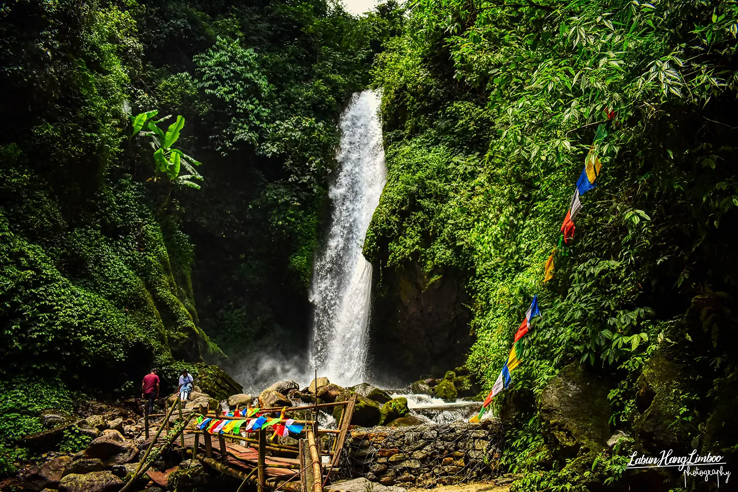 Cascading Kanchenjunga Waterfalls in West Sikkim, featuring a wooden bridge and colorful prayer flags.