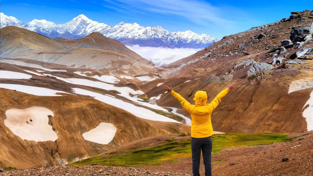 Trekker in a yellow jacket overlooks the dramatic snow-patched brown mountains and towering Himalayan peaks of Sikkim under a clear blue sky, showcasing Sikkim’s natural beauty and ideal trekking destination.