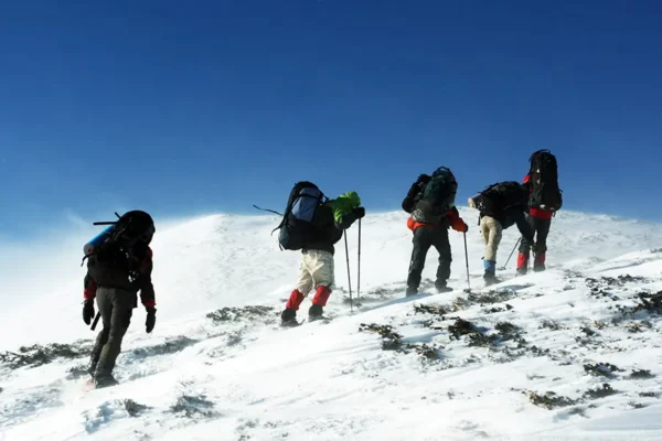 Team of mountaineers with heavy backpacks ascending a steep, wind-blown snowy ridge under a blue sky.