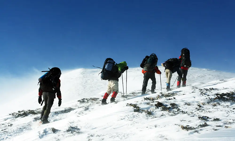 Team of mountaineers with heavy backpacks ascending a steep, wind-blown snowy ridge under a blue sky.